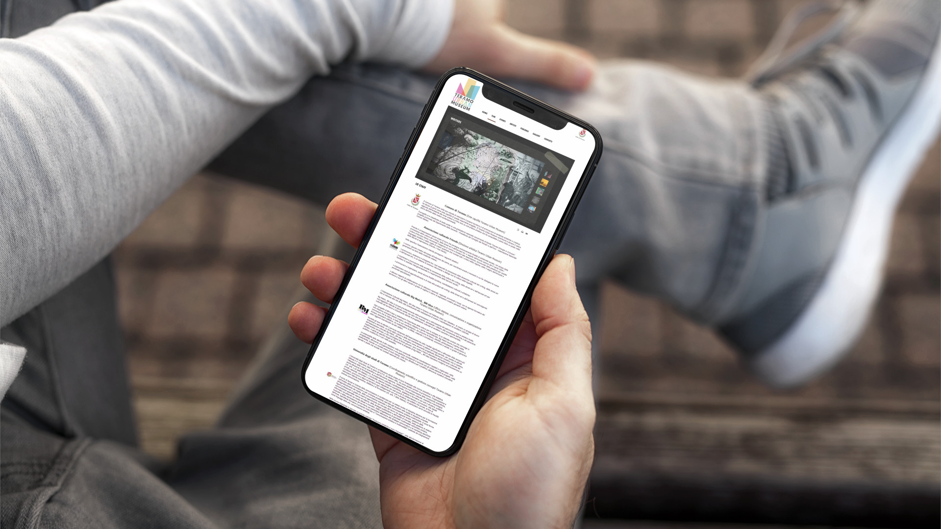 Mano che regge iPhone che mostra il sito web in modalità mobile responsive di Teramo Urban Museum