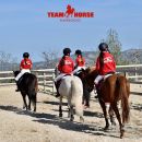 Tre allievi Team Horse in divisa rossa a cavallo nel campo del maneggio