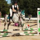 Amazzone con cavallo grigio durante il salto di un ostacolo verde